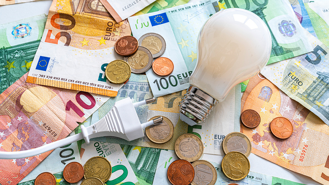 Eine Glühbirne, ein weißer Stecker und Münzen liegen auf verschiedenen Euro-Geldscheinen.